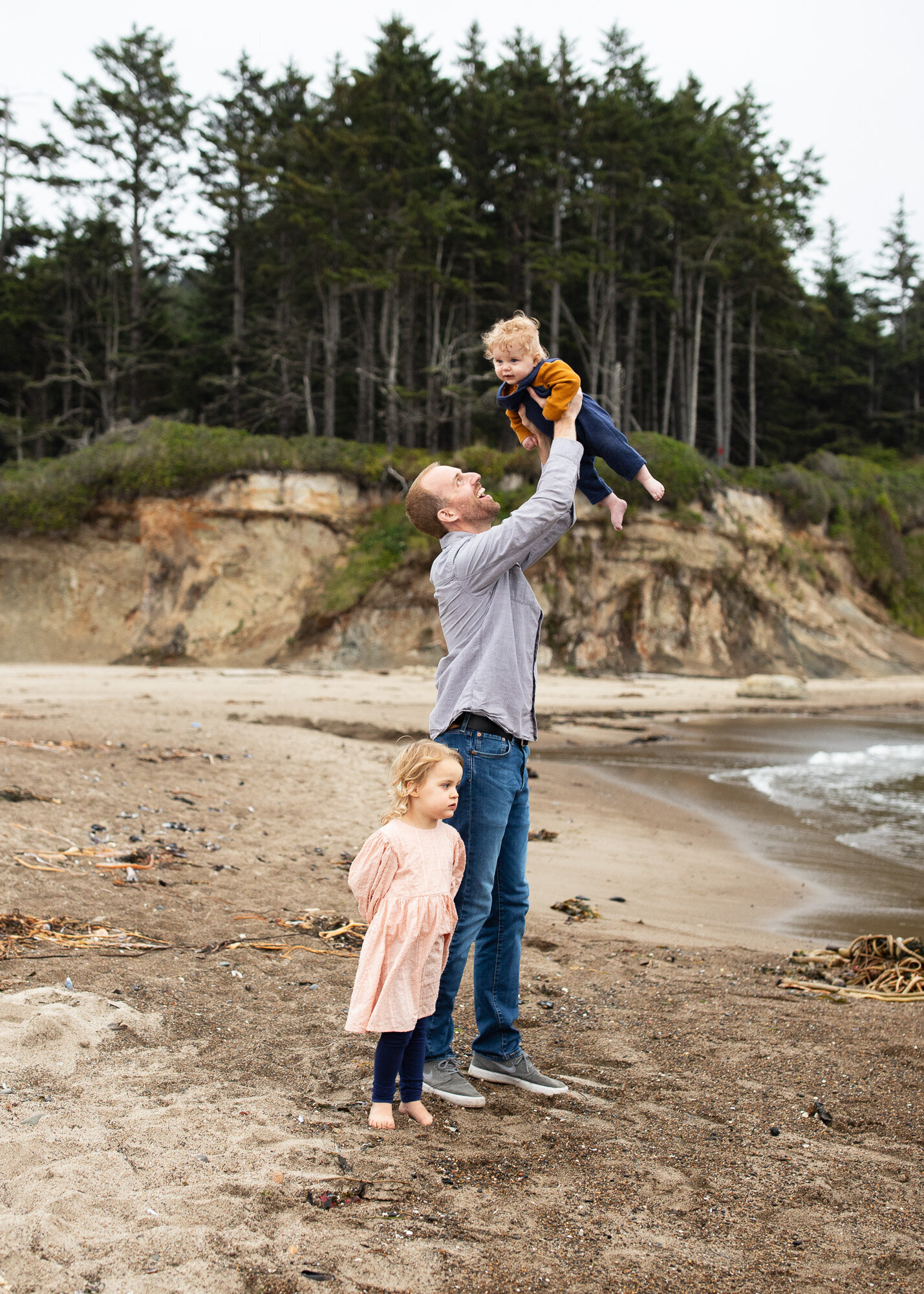 Dad holding baby above his head with toddler at his feet on Oregon beach. Photo by Oregon coast photographer.