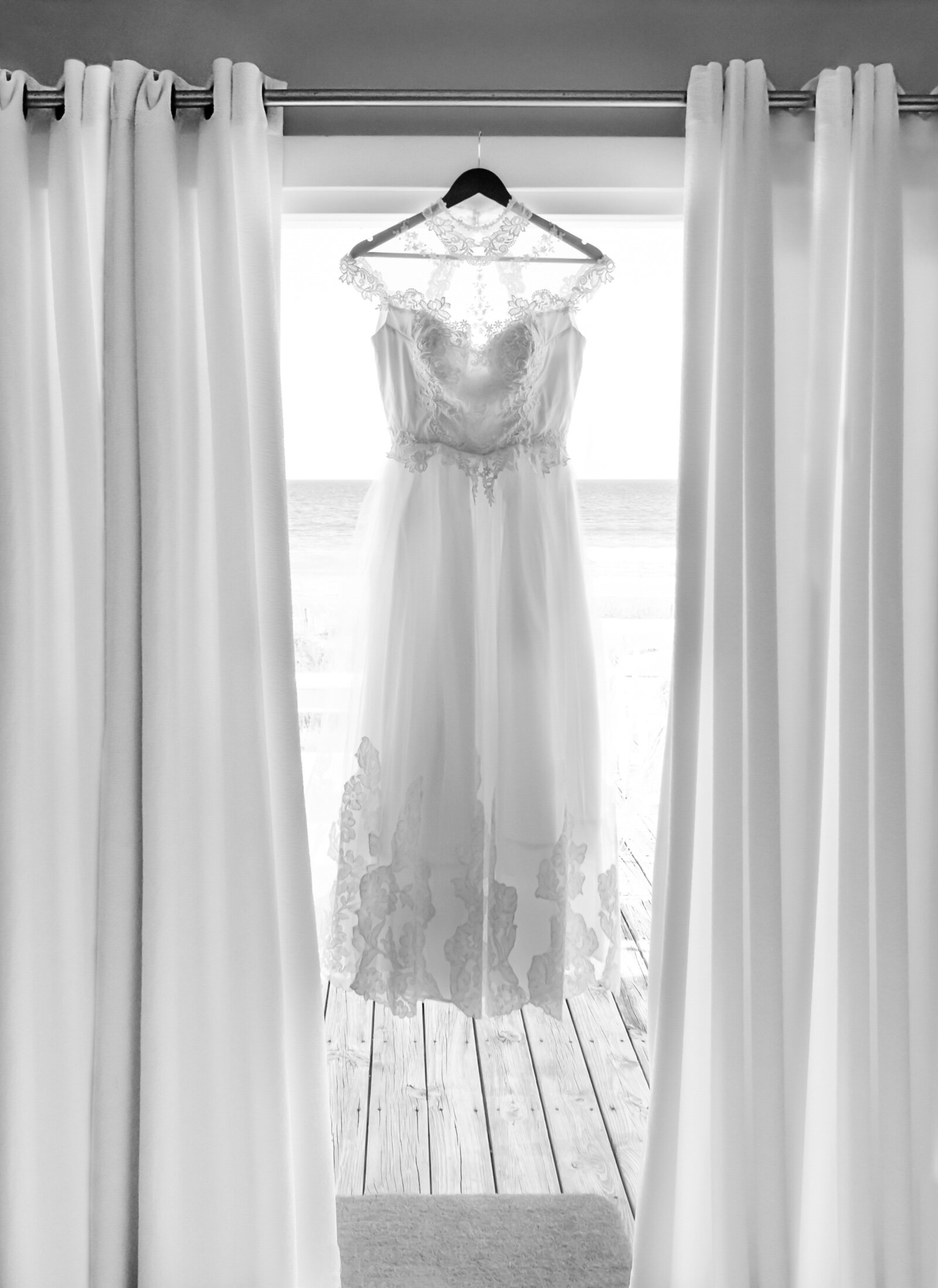 A breathtaking bride's wedding dress elegantly hanging in front of sheer curtains, with ocean views in the background, capturing a serene and romantic atmosphere in Wrightsville Beach, NC.