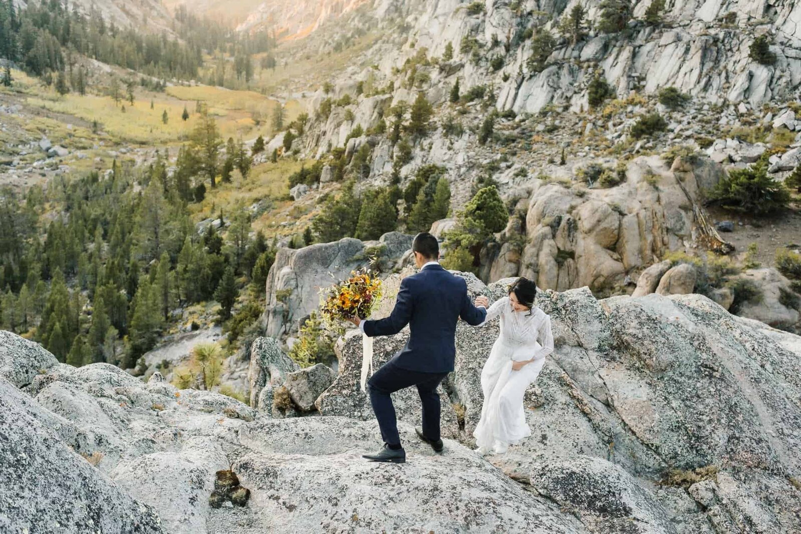 A very adventurous bride and groom climb up the side of a rock face overlooking a beautiful mountain valley on their mountain wedding day. 