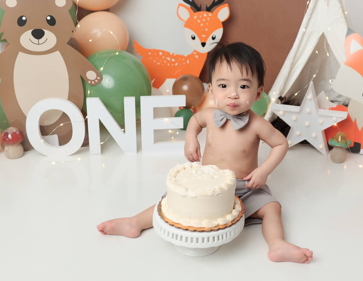 A handsome boy celebrating his first birthday with a woodland themed cake smash in a Rochester, NY studio. The scene features a beautifully decorated white cake surrounded by nature-inspired decorations, capturing the playful essence of the moment.