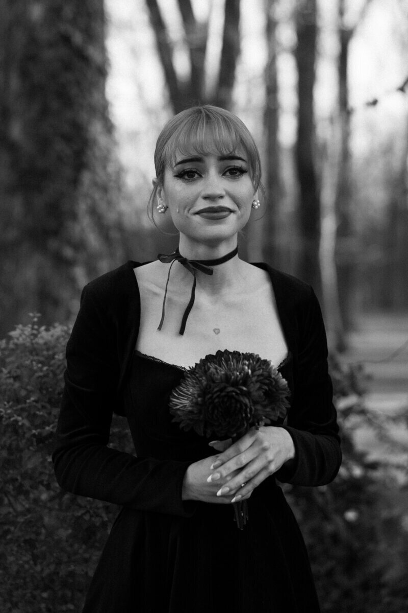A person stands outdoors, holding a bouquet of flowers. They are wearing a black dress and a choker necklace. The background features blurred trees and foliage. The image is in black and white.