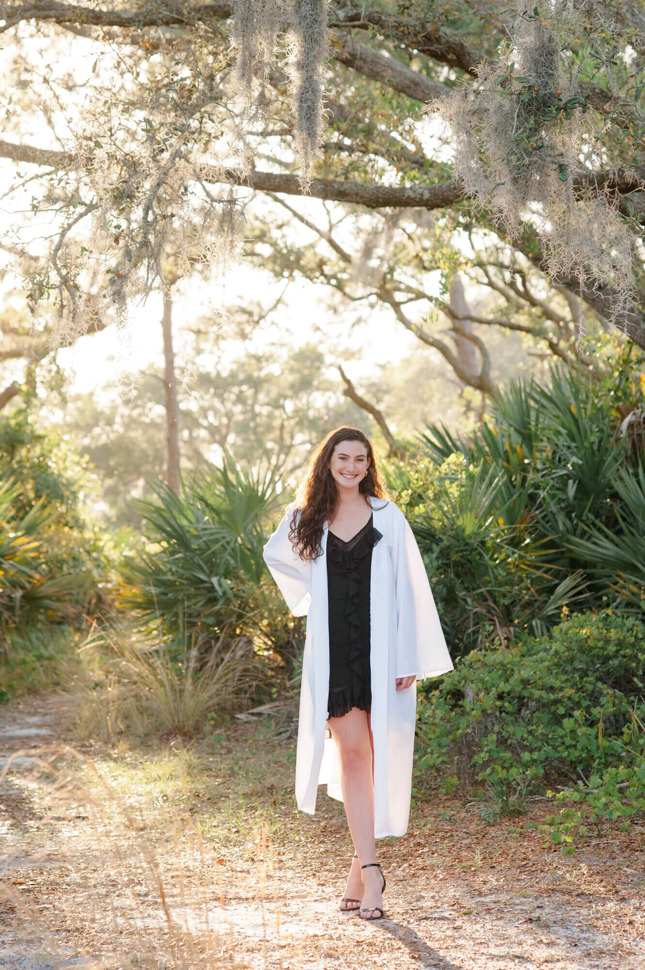 Senior girl wearing her gown stands next to a tall mossy oak tree during her senior photoshoot at WIckham Park