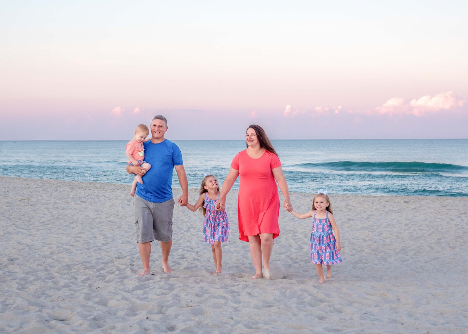 A family of five walks hand in hand along Carolina Beach at sunset, dressed in vibrant summer attire. The pastel sky and gentle waves create a serene backdrop. Perfect for artistic family photography in Wilmington, NC, highlighting joyful family moments.