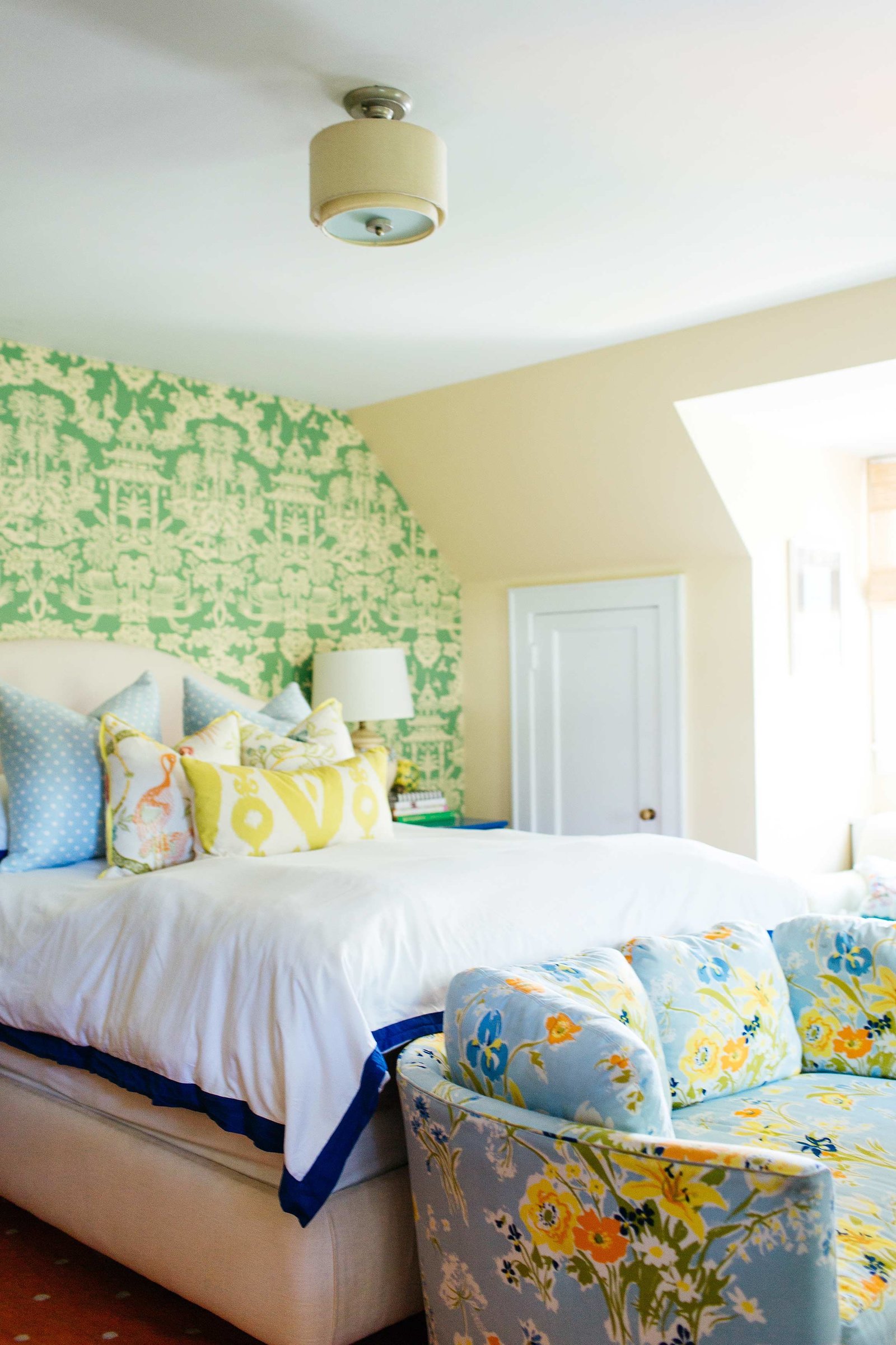 A white bed and floral love seat in front of a green wall papered wall.