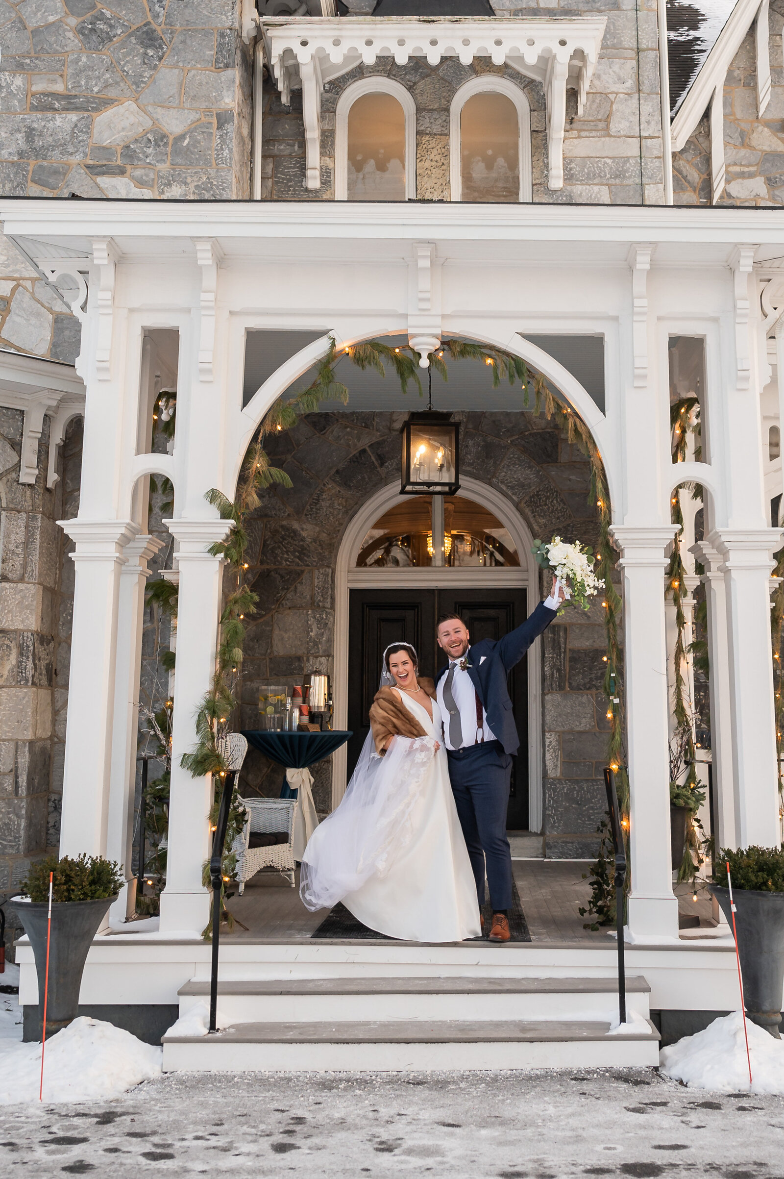 Bride and groom standing on the porch of an old stone building with the groom holding a bouquet of flowers in the air
