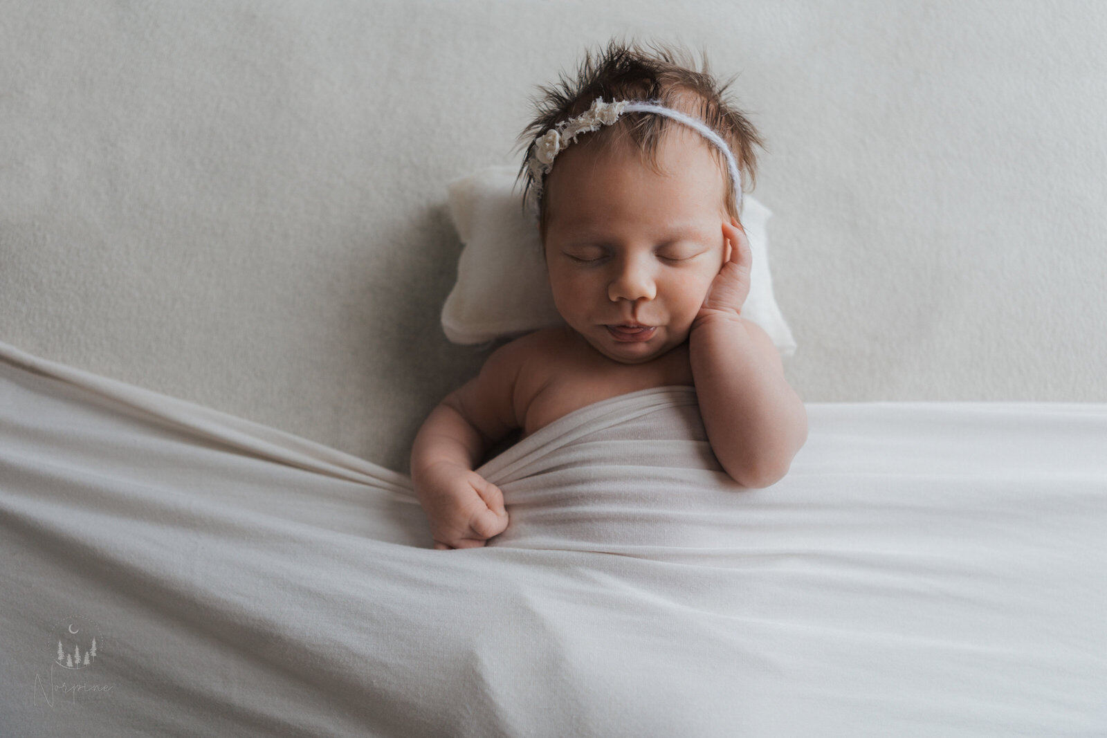 a newborn girl in the tucked in pose with a white background, blanket, and pillow, wearing a white headband. She is holding her face with one hand, and pursing her lips with eyes closed.
