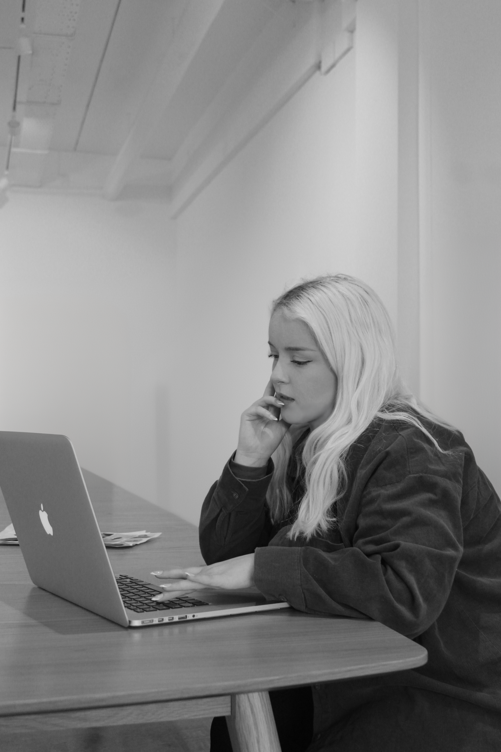 A black and white image of a woman sitting at a desk, her hand is to her lips, the other is scrolling on her laptop.