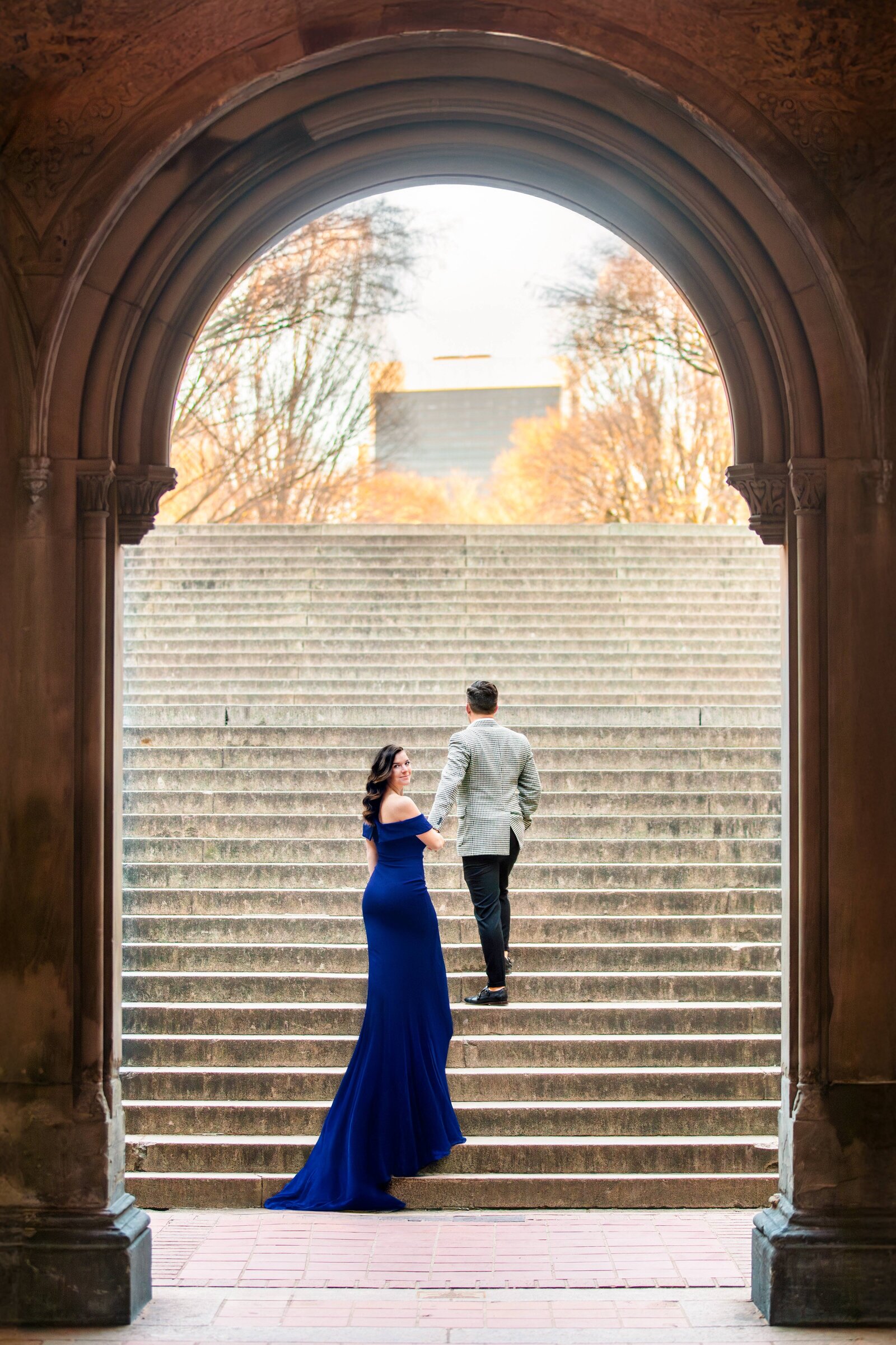 A woman in a long blue dress and a man in a gray suit walk up a grand staircase framed by an archway. The background features trees with golden leaves, suggesting an autumn setting.