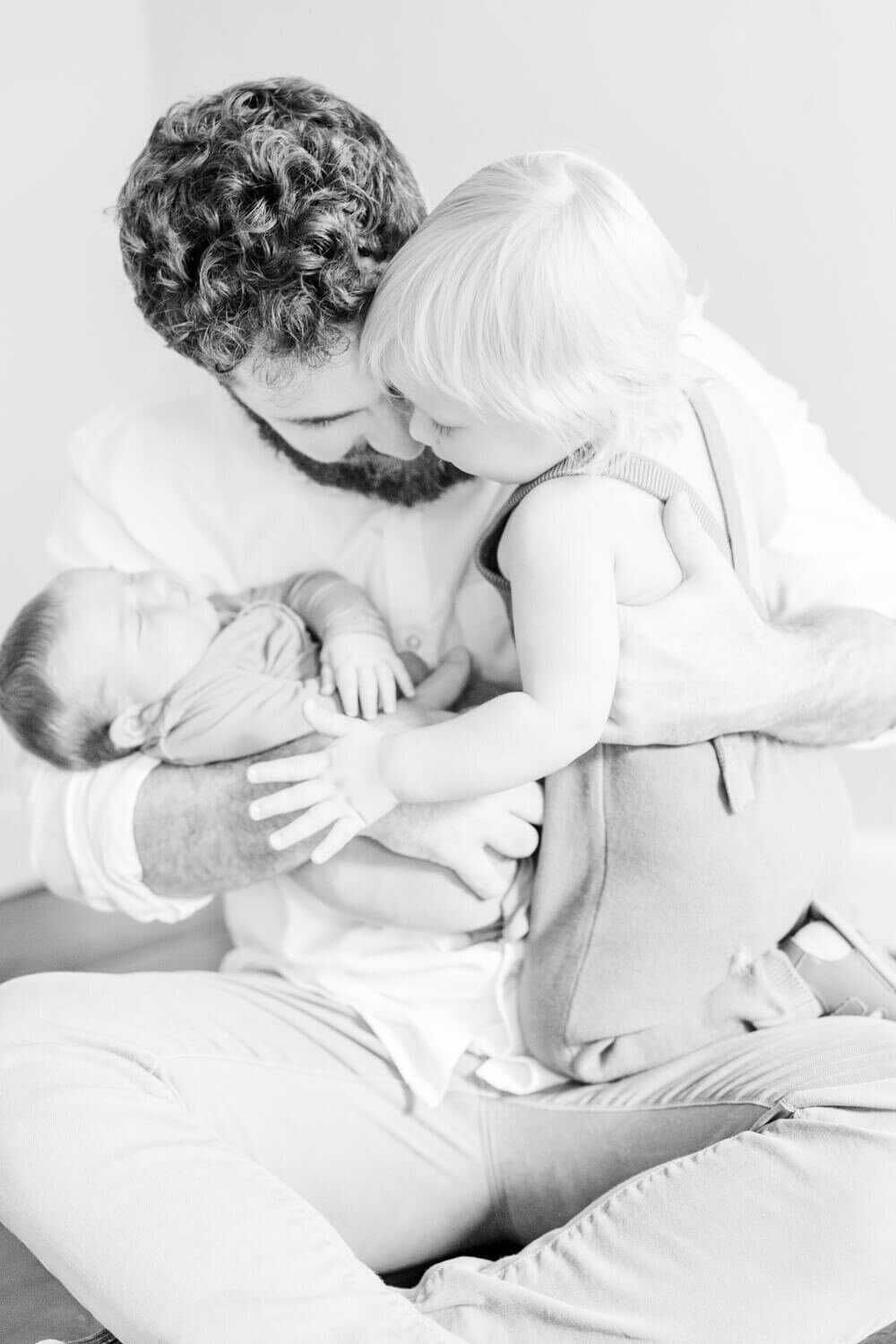 Dad holds newborn in one arm and his toddler in the other and the toddler watches the baby