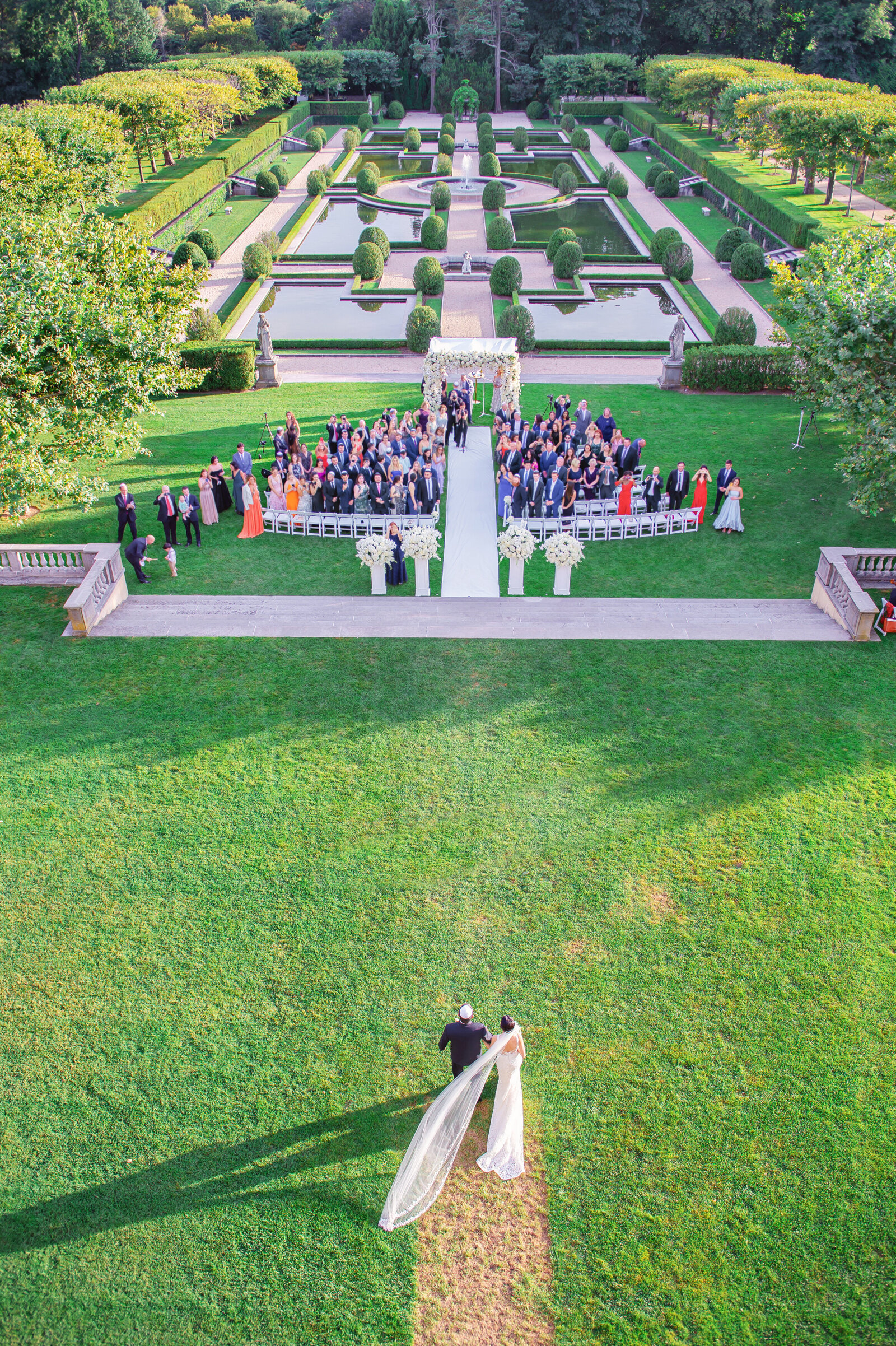 Aerial photo of bride walking towards wedding ceremony outside in a manicured garden
