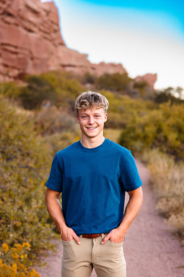 Senior boy in blue shirt puts hands in pockets while standing on a trail at South Valley Park in Littleton, Colorado.