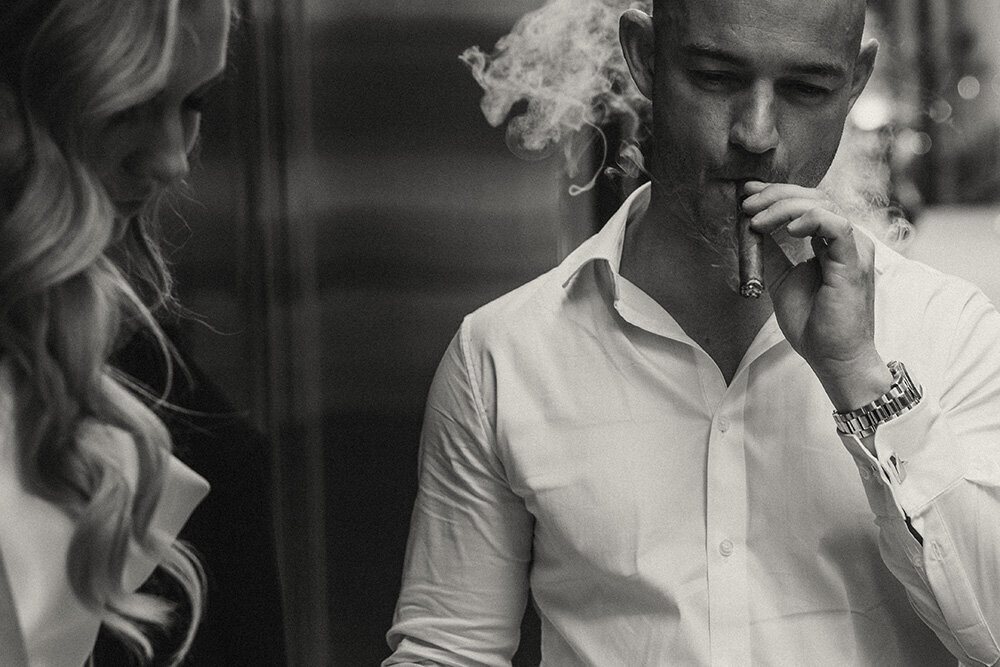 Black and white portrait of a groom celebrating elopement with a cigar with his bride close by.