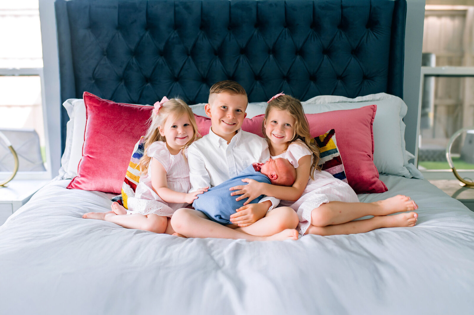 Parents cuddling their newborn and young children on a bed during a cozy in-home newborn session in Allen, Texas, photographed by Jennifer L. Kirk Photography.