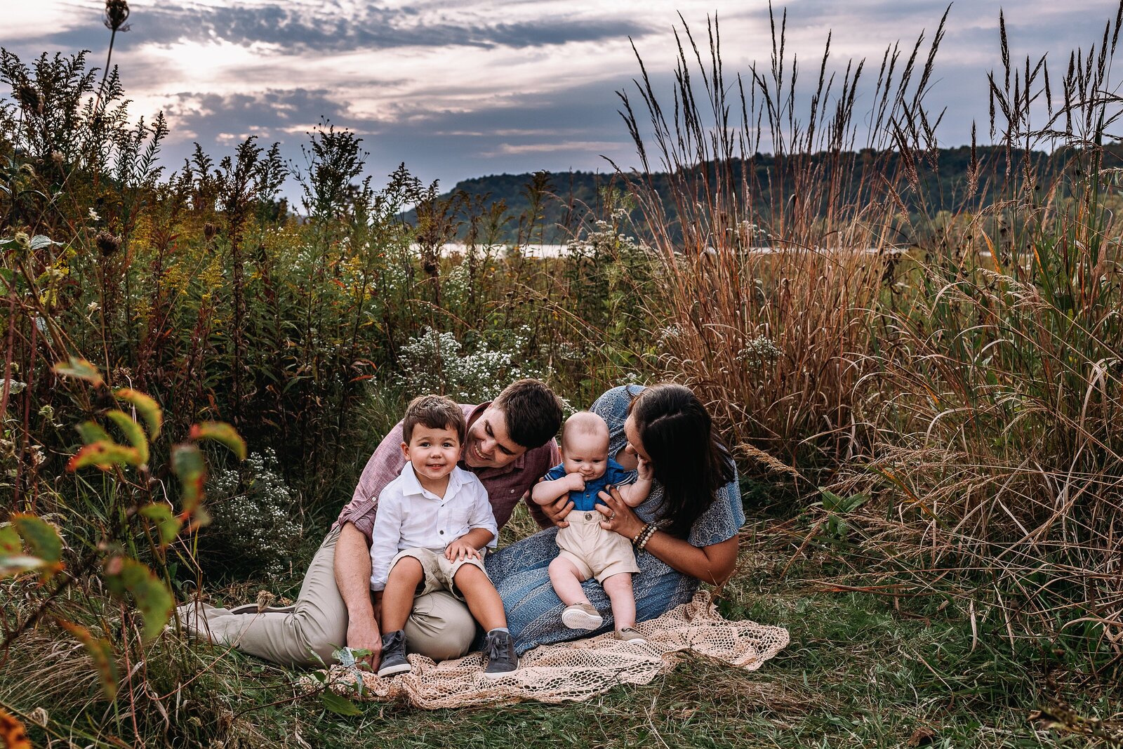Parents sitting on lace blanket holding toddler and baby in a tall field of grass and flowers, sunset and lake behind them.