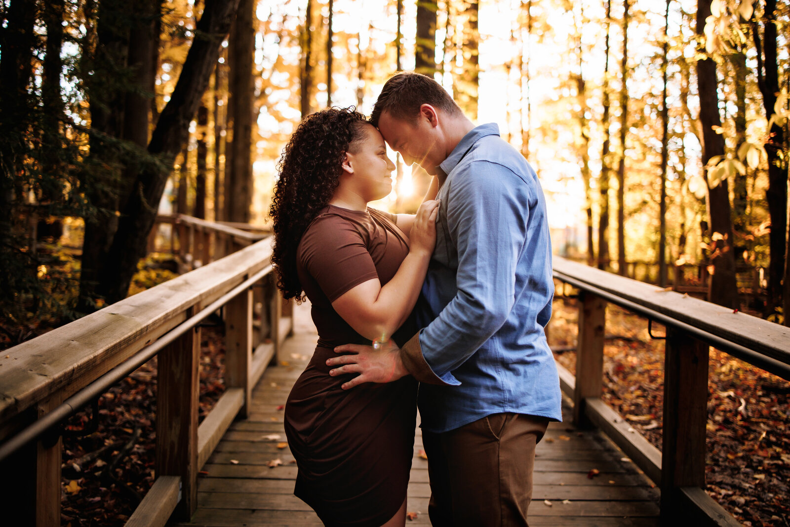 Couple stands with their foreheads together along a wooden walkway  with the golden sun peering through them
