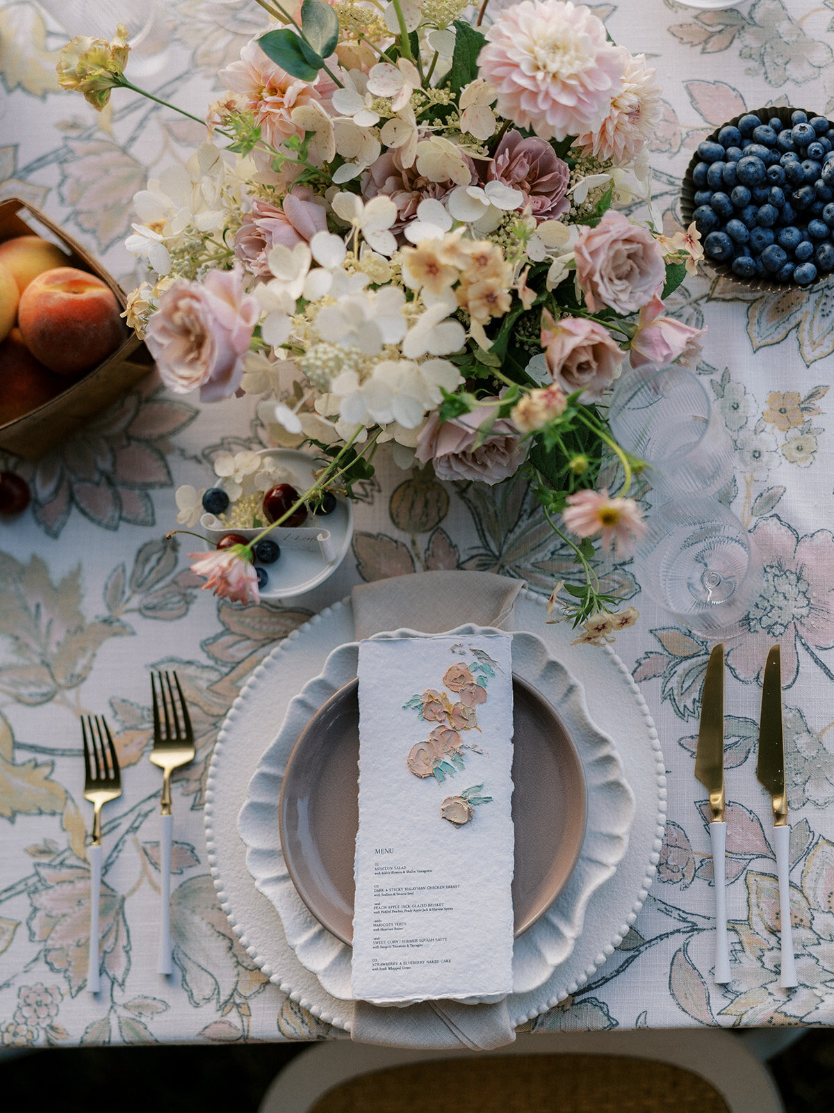 Flat lay photo of table setting on top of a floral-patterned linen, with fresh fruit and wispy floral compotes going down the length of the table including taupe and blush garden roses, white hydrangea, brown lisianthus, phlox and blush cosmos.