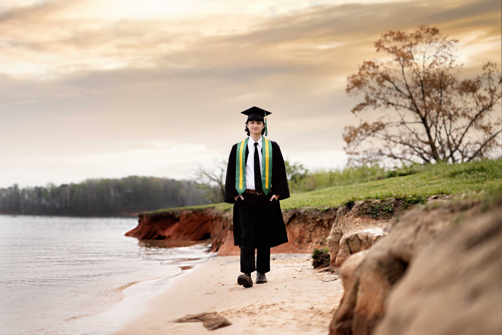Graduation photo of boy in cap and gown, walking on lake and looking into the camera smiling. Senior graduation photos taken by Jenni Nikki Photography in Atlanta, GA