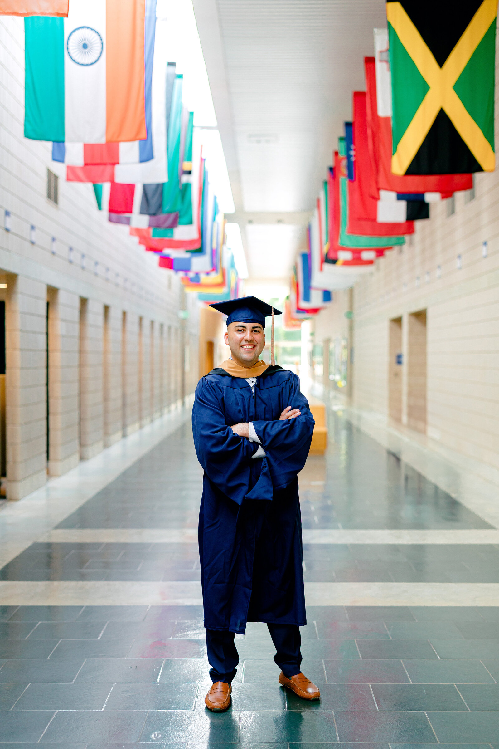 Duke MBA graduate wearing cap and gown smiling for portraits inside Fuqua School of Business with international flags hanging above.