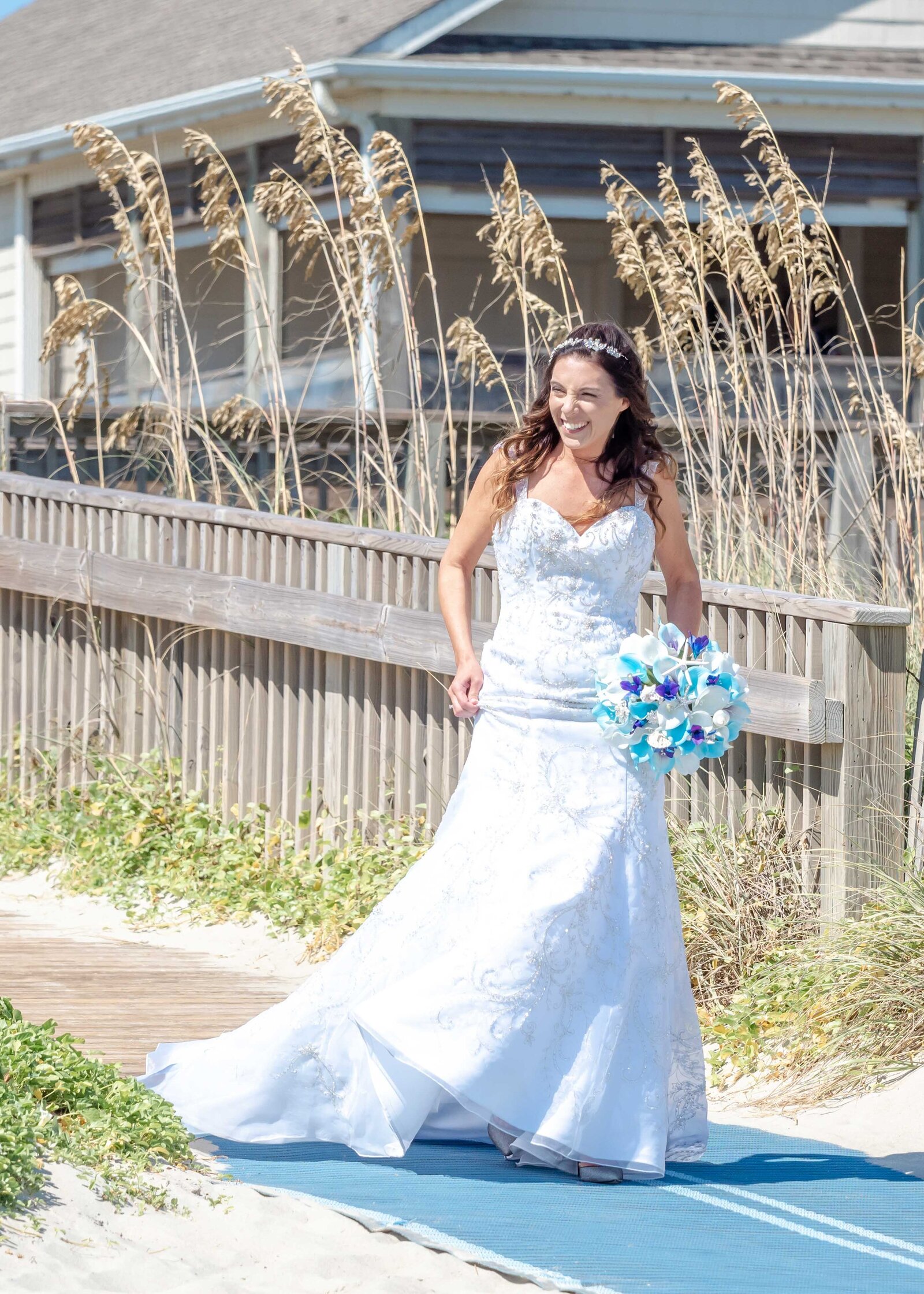 A smiling bride in a lace gown carries a vibrant blue bouquet while walking to her beach ceremony, captured at a wedding in Topsail Beach, NC, blending coastal charm and authentic moments.