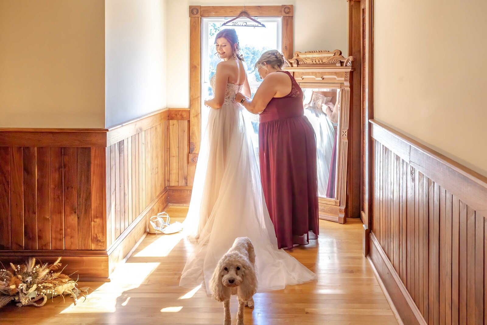 A bride gets ready with the help of her mother in a rustic bridal suite, while her small dog waits nearby, captured during a Wilmington, NC wedding, showcasing authentic moments and pet-friendly charm.