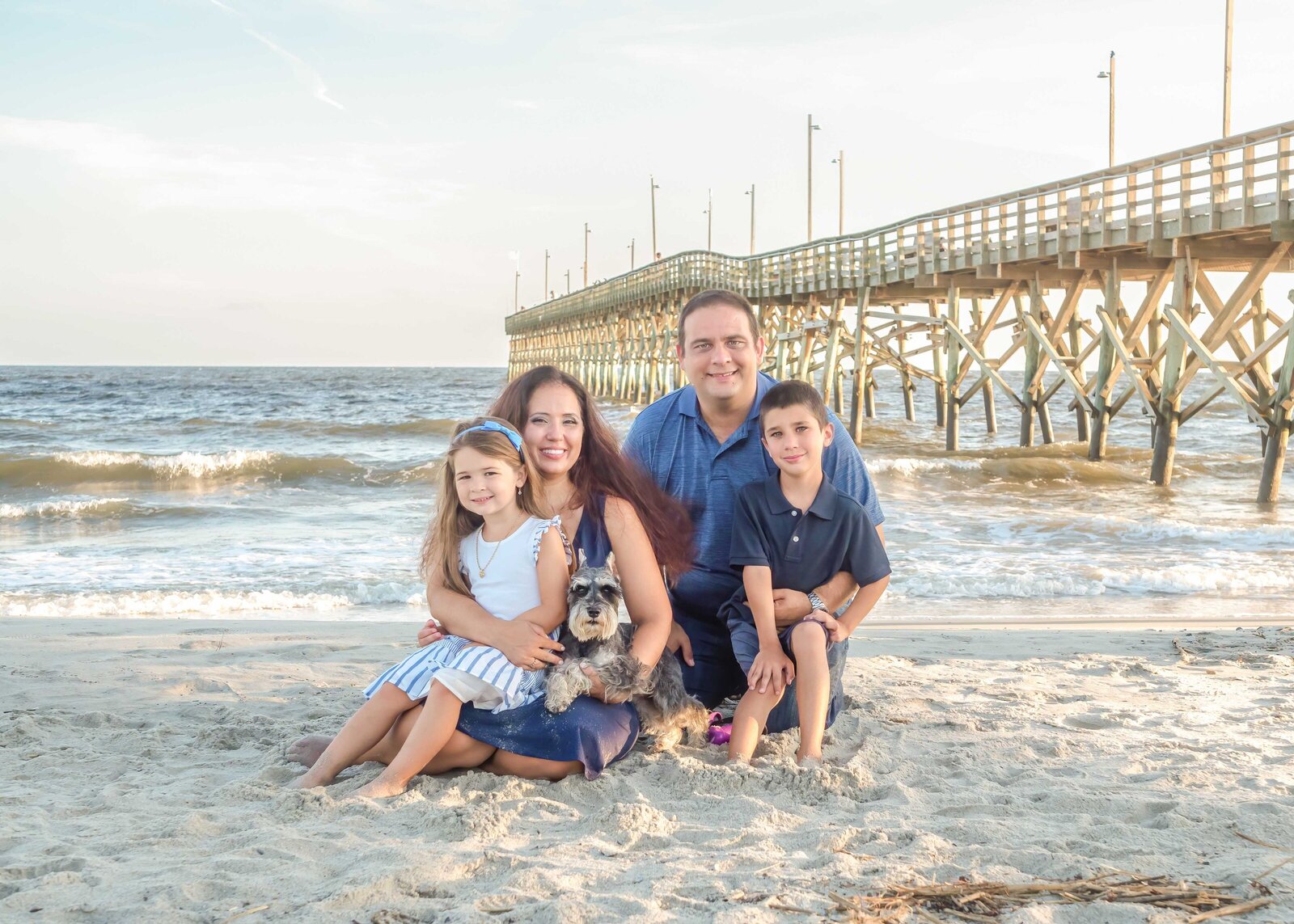 A family sits with their dog on the sandy beach near a wooden pier, dressed in coordinated blue and white outfits. The ocean waves and pier create a picturesque setting. Ideal for affordable family photography in Wilmington, NC, highlighting cherished family moments.