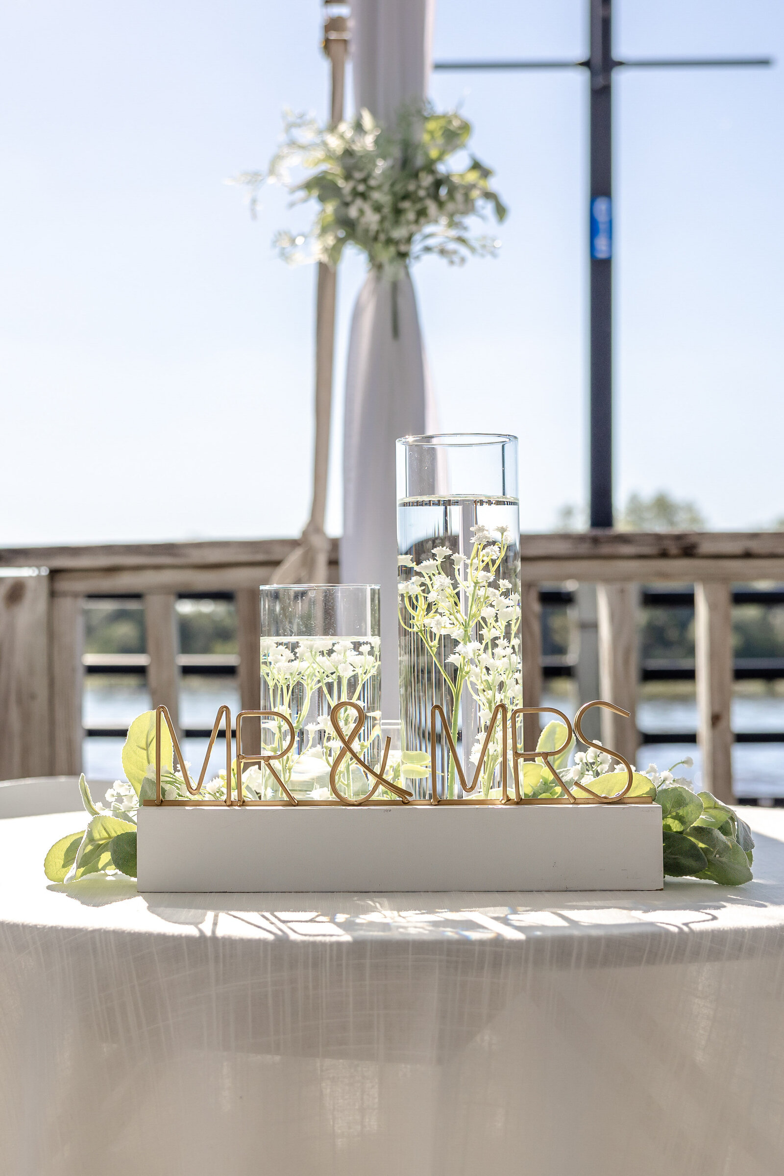 A unity ceremony table featuring floating candles, greenery, and a gold "Mr. & Mrs." sign, captured during a Wilmington, NC wedding on a waterfront deck, blending elegance and coastal charm.