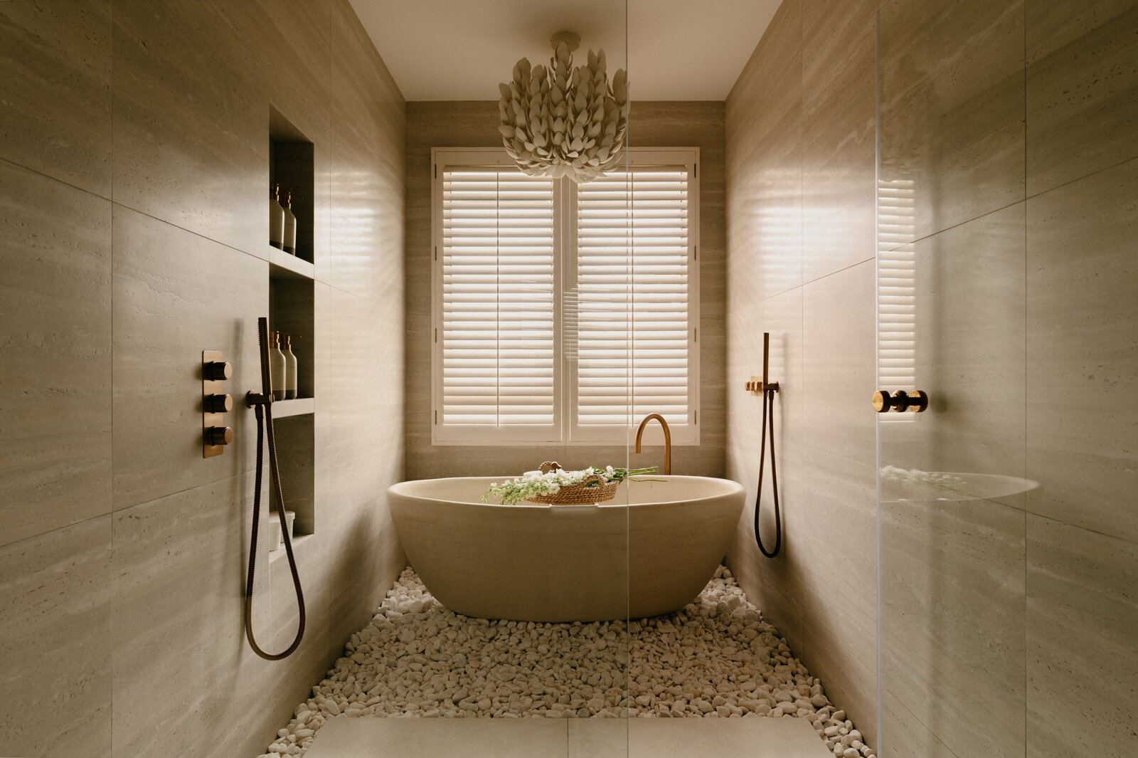 Freestanding stone bathtub set on white pebbles with brushed brass fittings, framed by natural stone walls and shutters.