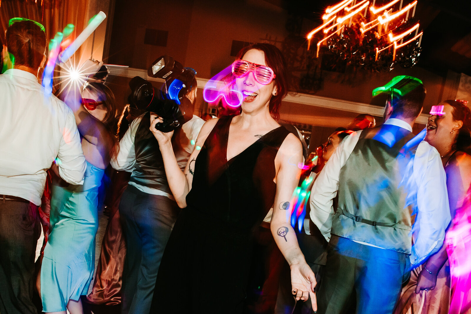 Documentary-style New England wedding photographer capturing reception moments while dancing with guests, wearing light-up glasses and holding a camera