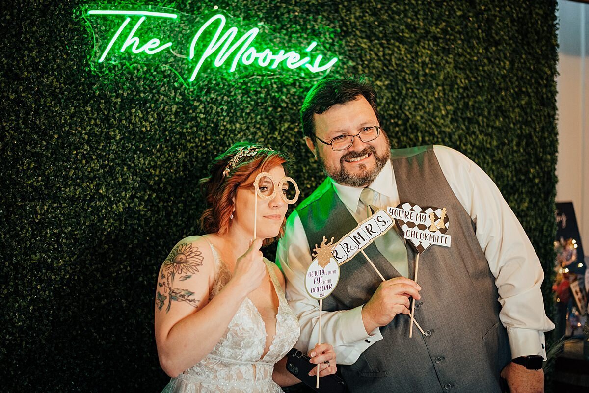 The bride and groom pose with their custom photo booth props and neon sign proclaiming them to be newlyweds