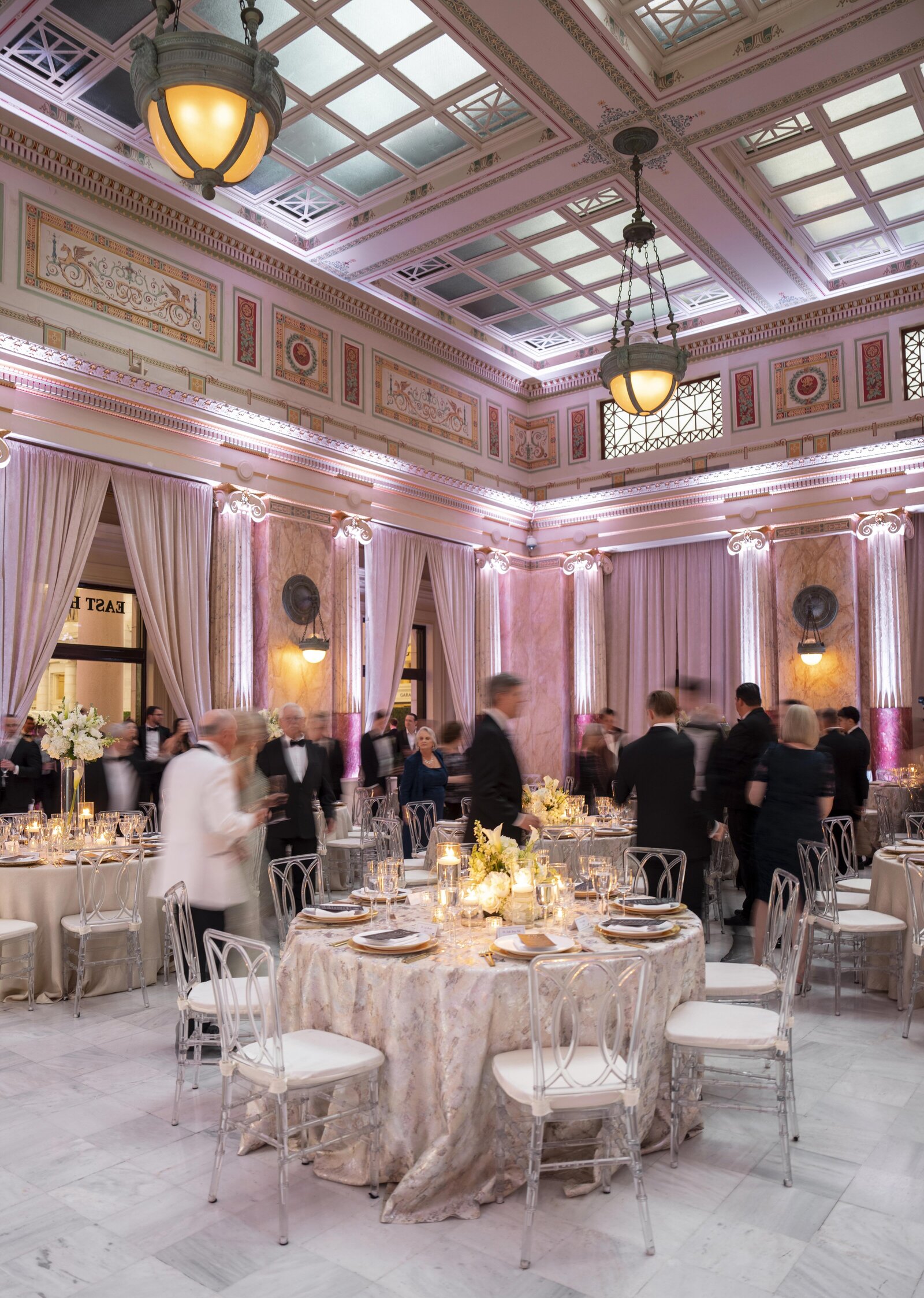 A grand ballroom with ornate ceilings and chandeliers, set for a formal event. Elegant tables are adorned with white and gold linens, floral centerpieces, and candles. Guests in formal attire are mingling around the tables.