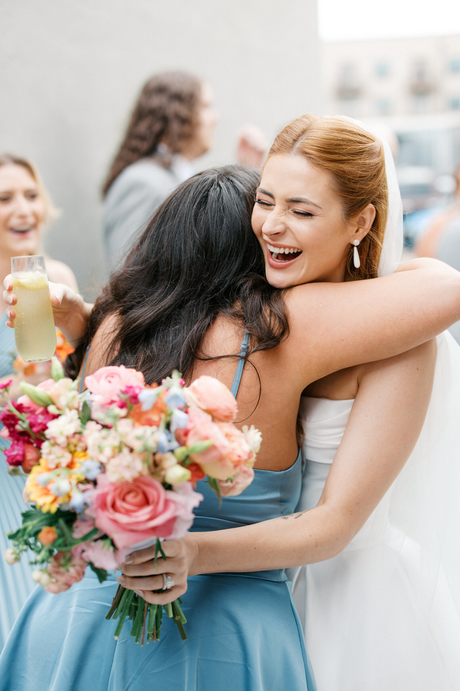 Red head bride embraces bridesmaid with colorful bouquet smiling from ear to ear and golding yellow cocktail