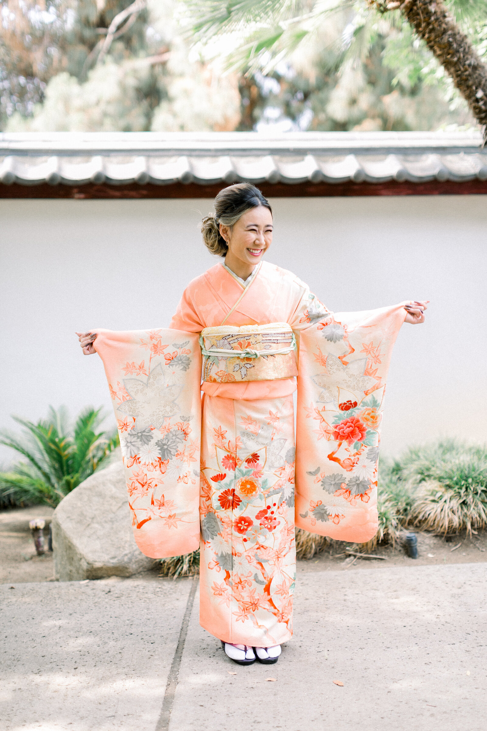 Full-body view of a young woman in a peach furisode at Brand Park tea house Glendale for her Seijinshiki coming-of-age celebration. Kimono rental in Los Angeles | ロサンゼルス・ブランドパーク日本庭園で成人式のために桃色の振袖を着た女性の全身ポートレート。着物レンタル