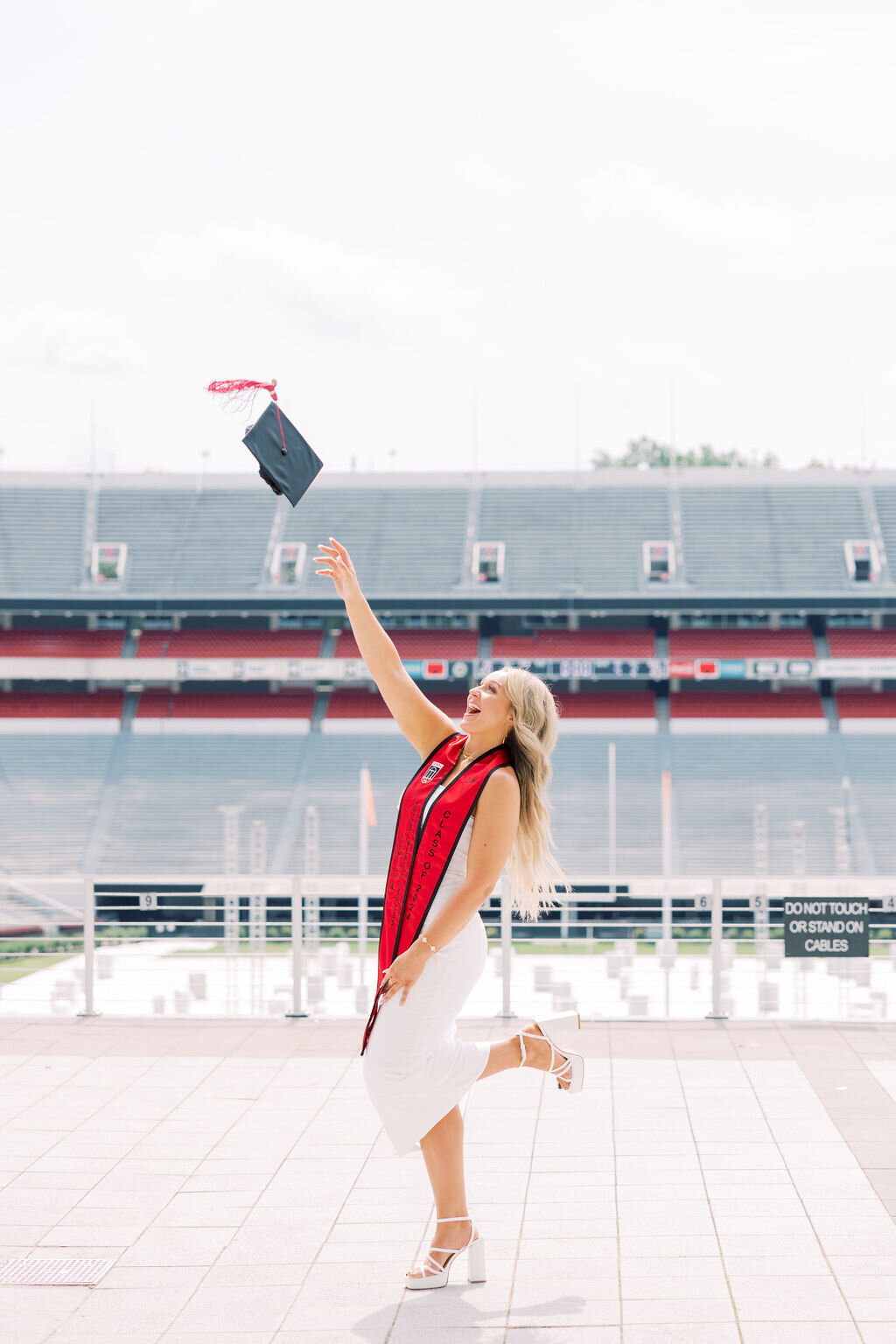 Girl in white dress and graduation stole throws cap at University of Georgia Stadium