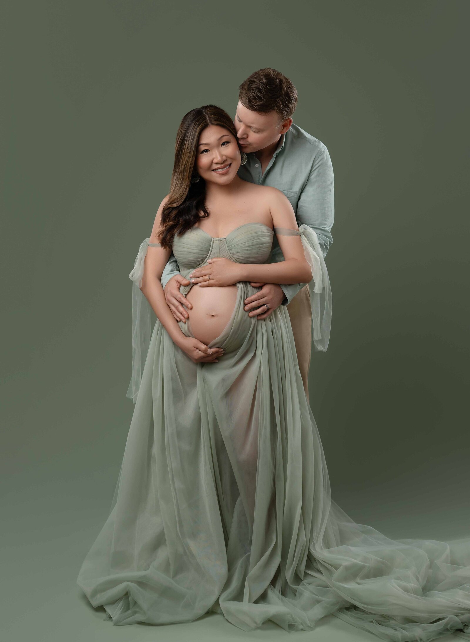 Dad kissing mom's cheek while snuggling and holding baby bump. Photographed at Atlanta photo studio.