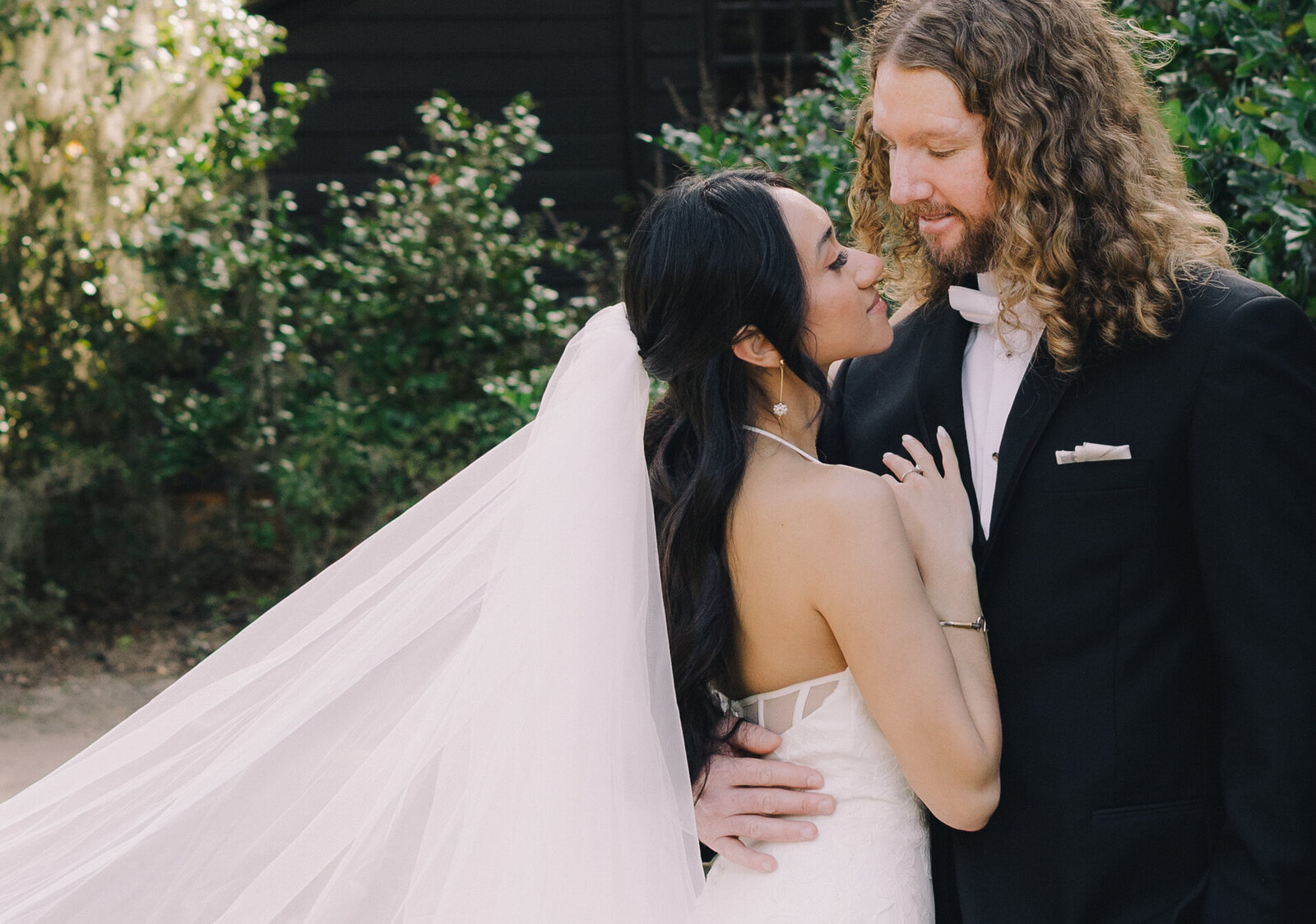 Romantic wedding portrait of a bride and groom embracing outdoors, with her long veil flowing behind her, captured in soft natural light by a luxury Charleston, South Carolina wedding photographer.