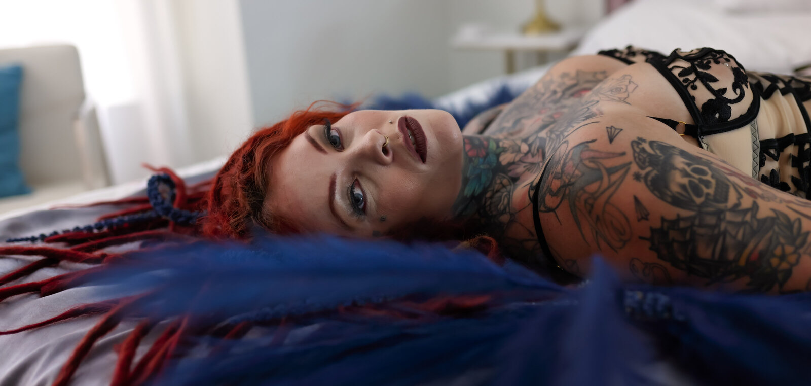 Savannah Georgia Boudoir Photography and Glamour boudoir portrait of Woman with tattoos and dreadlocks laying on bed looking in camera with feathers close up