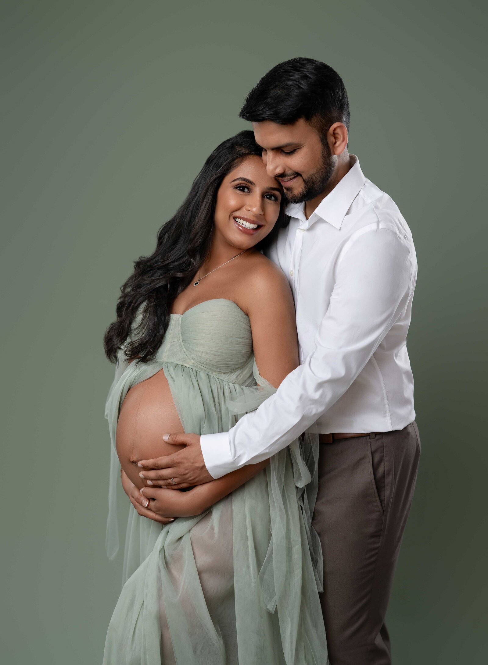 Mom and Dad cuddling as Dad holds baby bump and smiling. 
