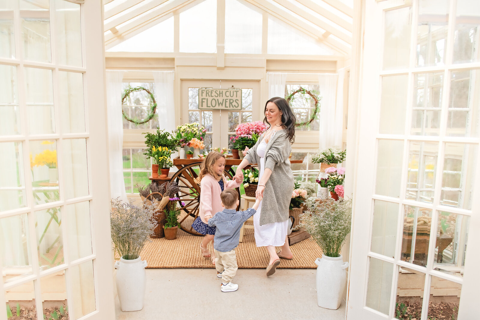 A mother dances joyfully with her children in a flower-filled greenhouse. Styled with rustic charm, this moment reflects the warmth and connection of family sessions by Meraki Photographic Creations in Dundas, Ontario at the All Who Wander Flower Farm.