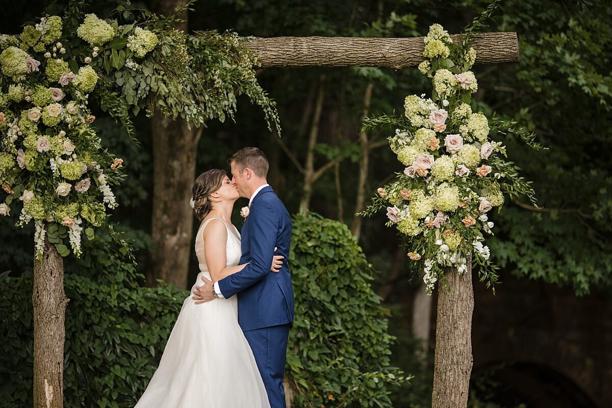 The groom, wearing a blue suit, kisses his new wife for the first time under a log arbor covered in white and blush flowers and greenery