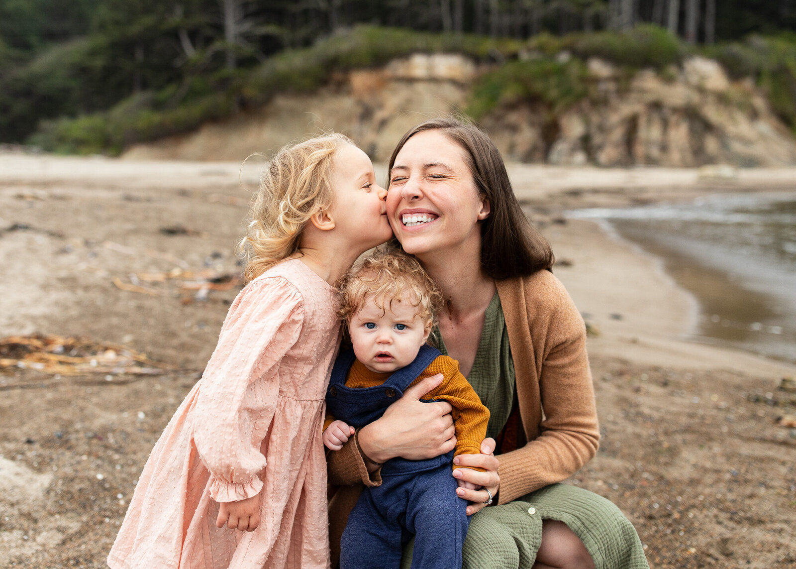 Mom kneeling in sand while holding her infant son and toddler daughter kisses her cheek. Oregon Coast Photographer