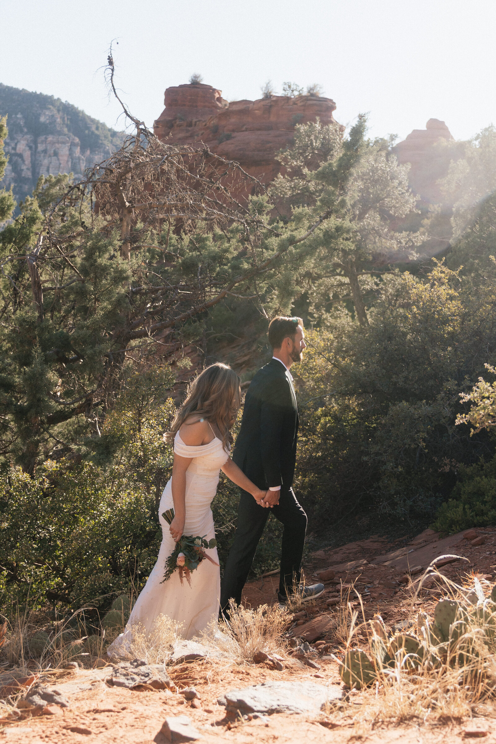 Bride and groom hiking through red rock trails and desert greenery during their adventurous elopement at Merry Go Round Rock in Sedona, Arizona.