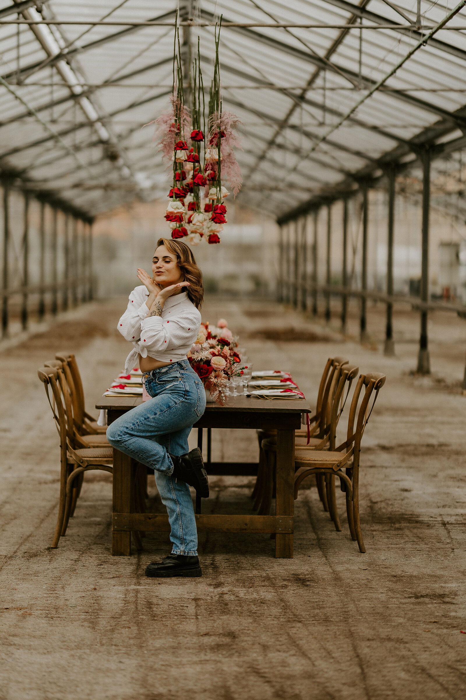 Laura, photographe de mariage en Vendée, fait une pose girly devant une table de mariage décorée, dans une immense verrière, avec des fleurs suspendues.