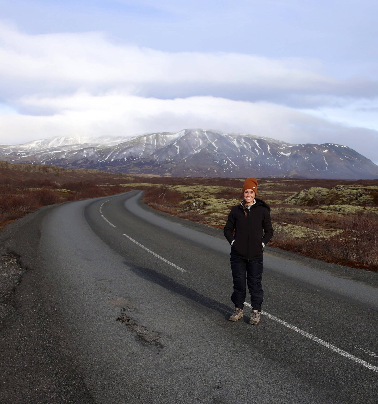 A smiling photographer stands in the middle of an empty rod smiling at the camera. There is green and red moss on the ground around here and a vast snow-topped mountain range in the background. 