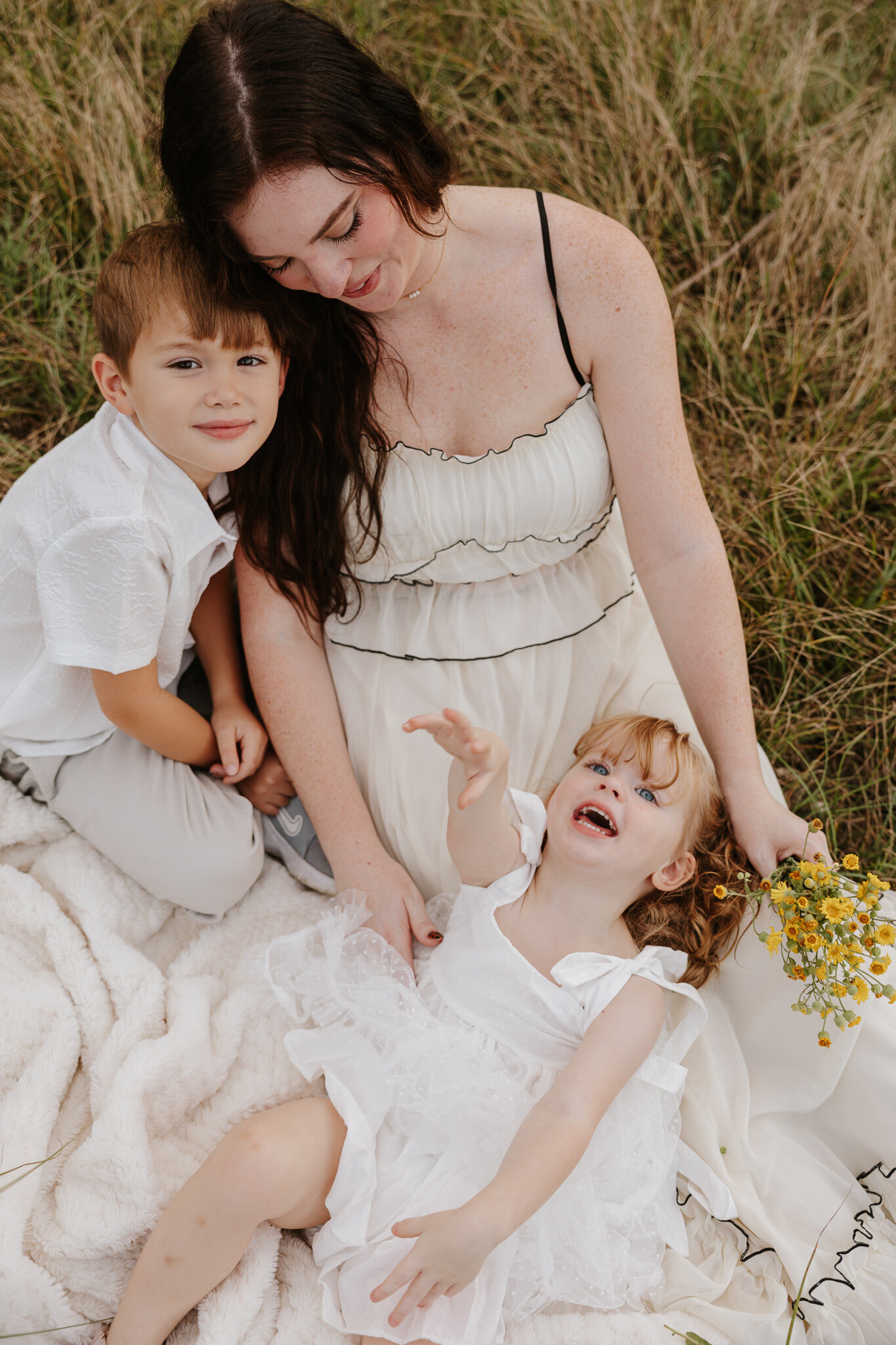 Mother sitting in farm field in Aiken SC with her two children - big brother smiling at the camera while the little sister laughs and holds wildflowers.