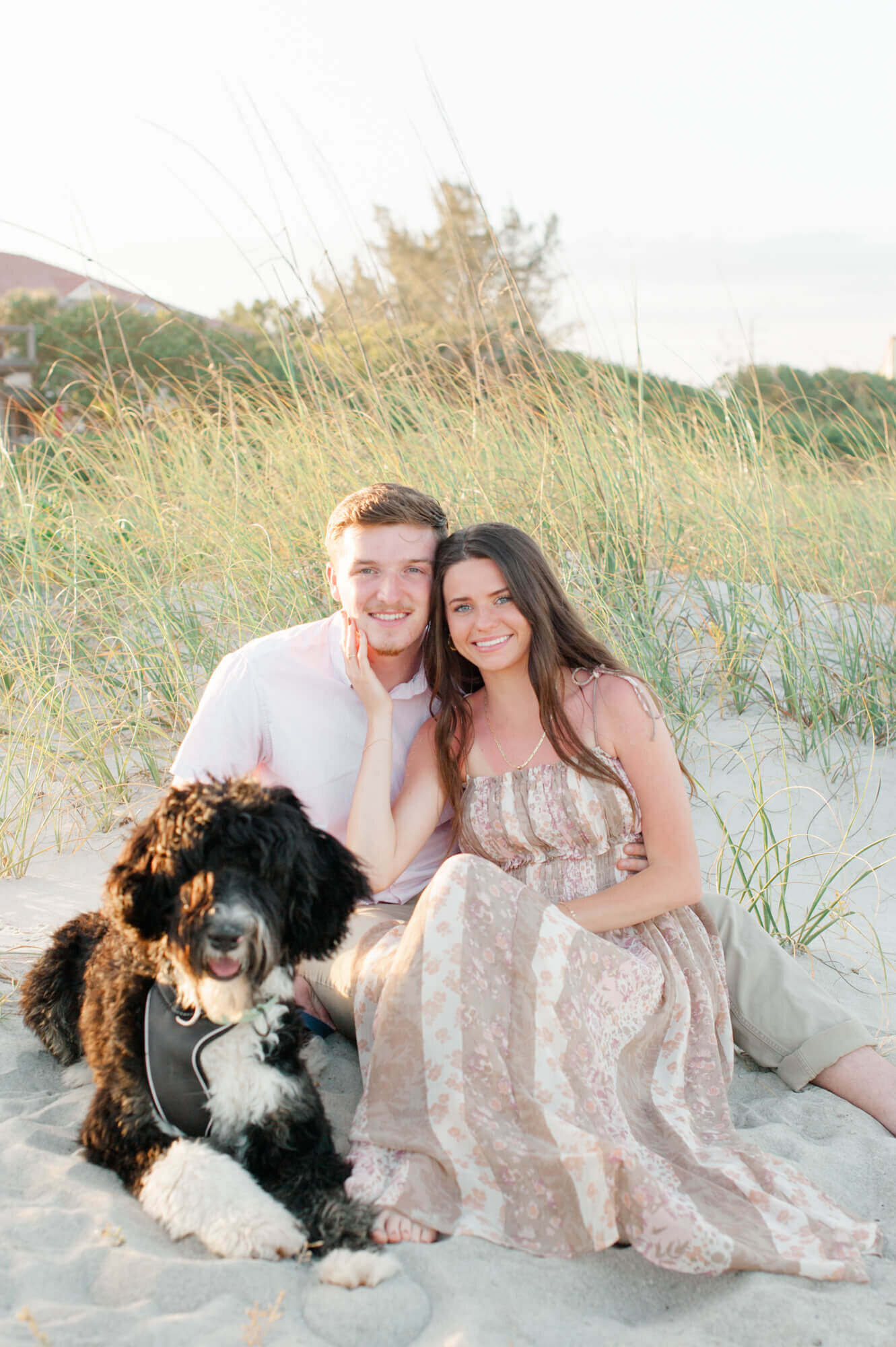 Couple holds their black dog and smiles at the camera sitting near the dunes for a portrait