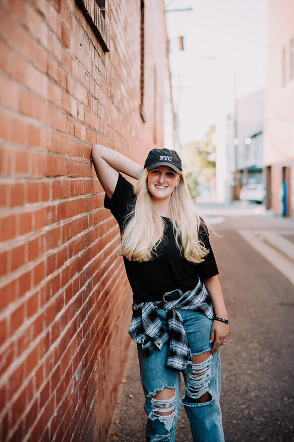 Girl in hat poses with arm up against a brick wall in downtown Littleton, Colorado.