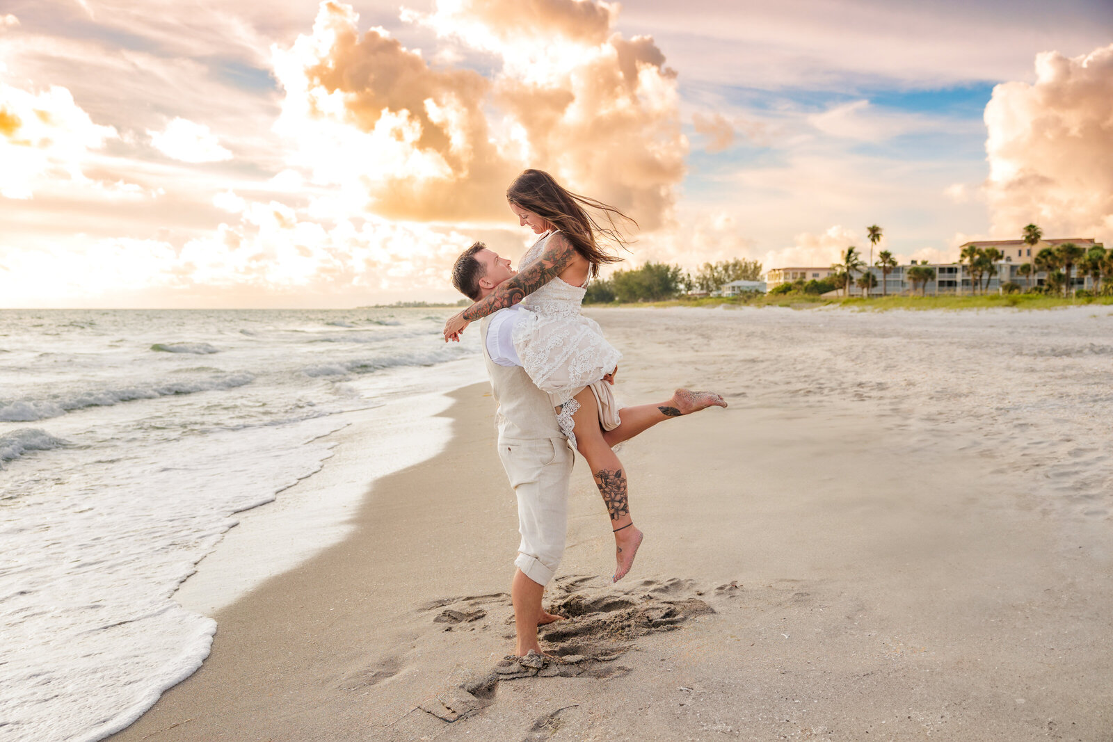 Bride and groom on the beach with the ocean to the left of them. The groom is lifting the bride, and she is looking down at him. The sun is setting in the background with buildings and houses to the right.