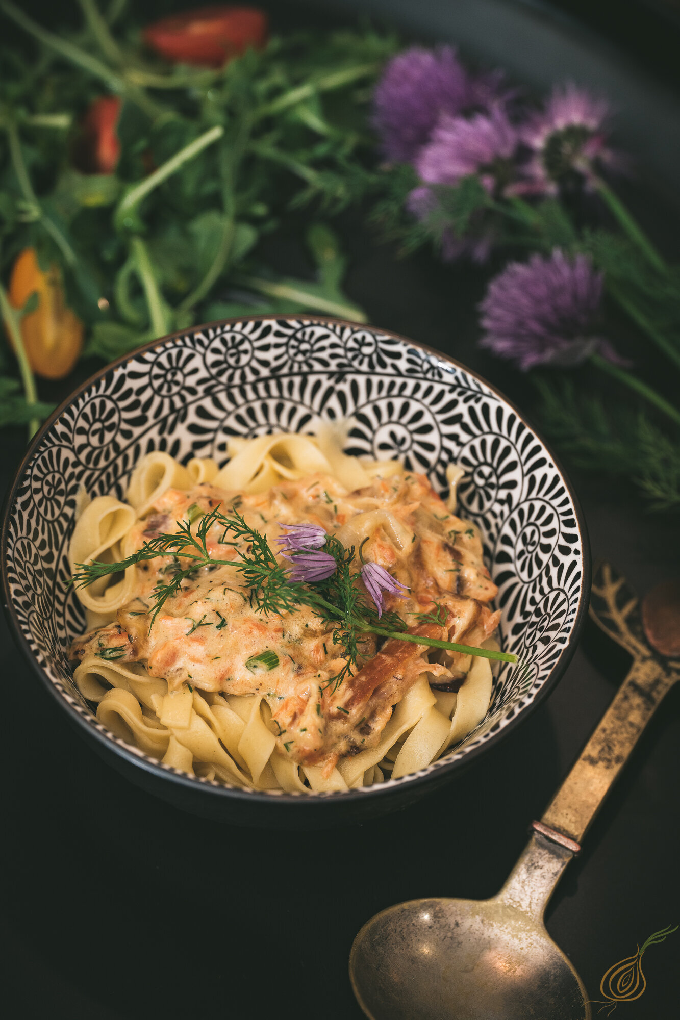 A bowl of salmon pasta garnished with fresh herbs and vibrant vegetables, beautifully arranged on a black table.