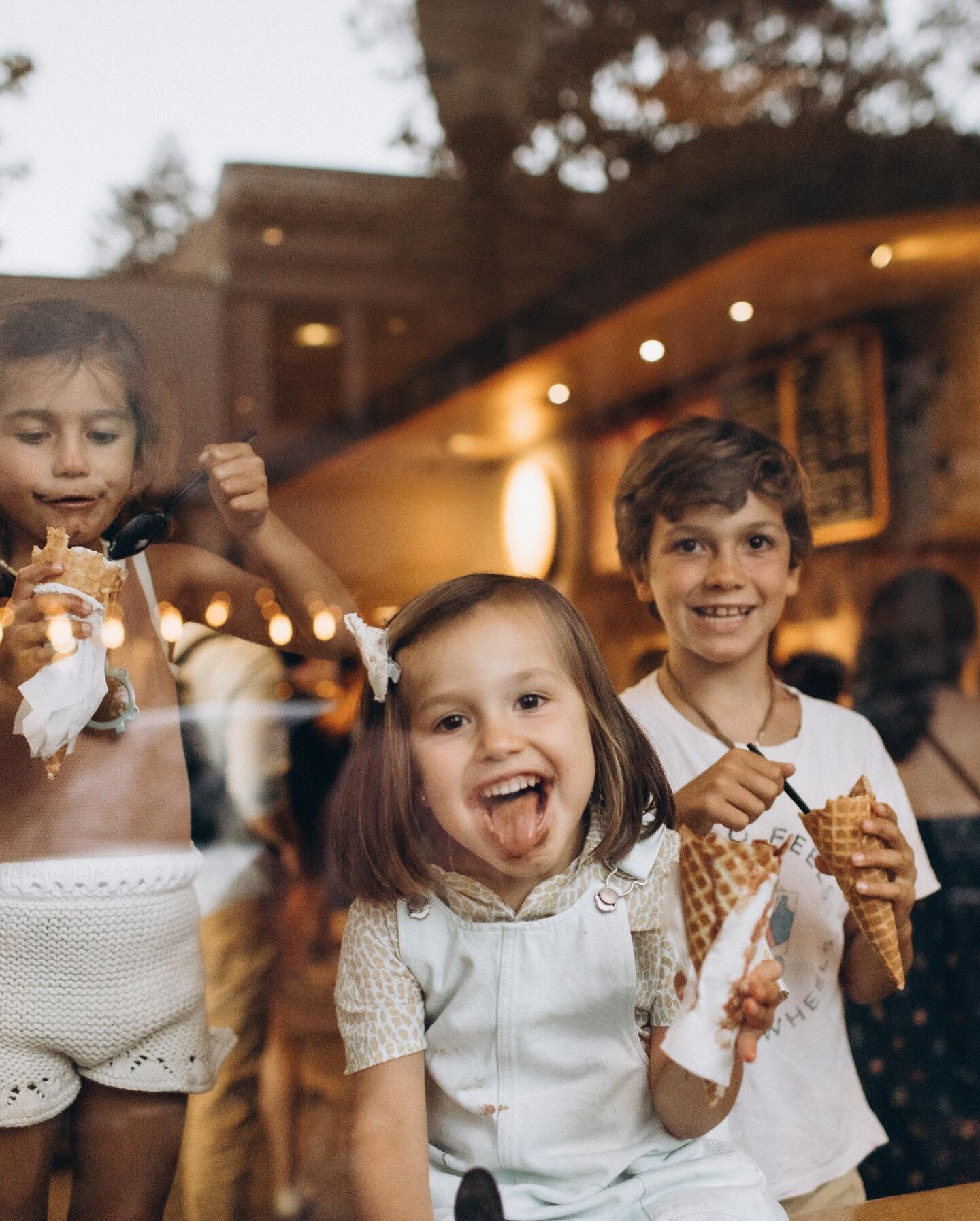 Three children eating ice cream; a girl with a big smile in the foreground, a boy behind her, and another girl to the left, all behind a glass window enjoying their ice cream.