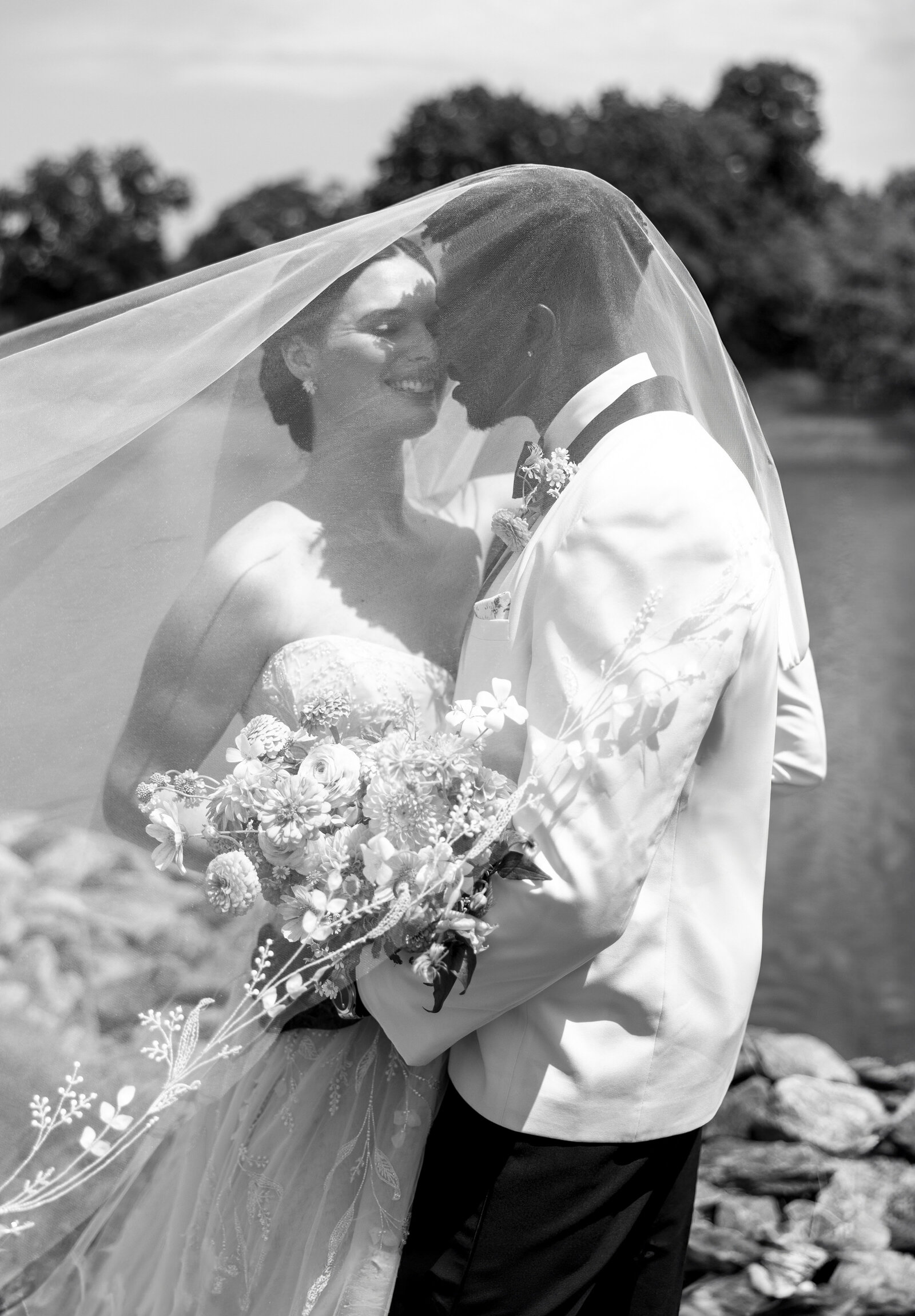 A bride and groom stand under a veil near a lake. The bride holds a bouquet of flowers, and both are smiling. The groom wears a tuxedo jacket, and the scene is set outdoors with trees in the background.